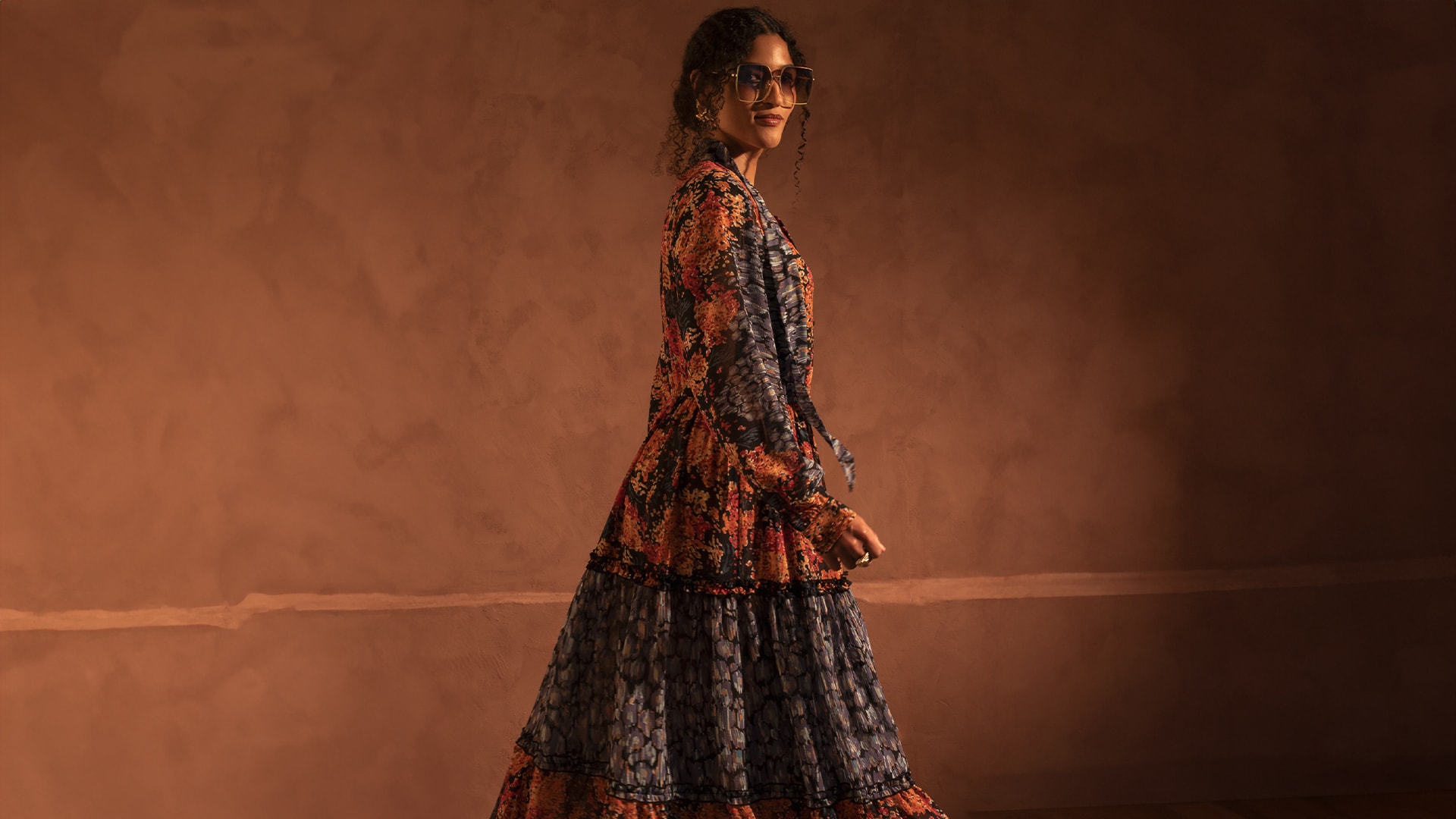 Person in a floral-patterned dress and sunglasses poses against a warm brown background, looking to the side with a confident expression.