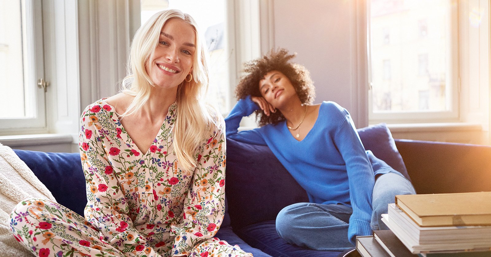 Two women sitting on a blue sofa. One woman is wearing a Indiskas floral dress 'Frida' and the other woman is wearing a bright blue knitted sweater. 
