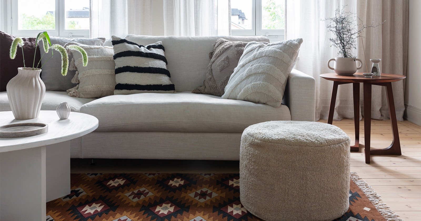 En stue med en hvit sofa med duskete puter, et fargerikt og mønstret teppe med en puff stående på det